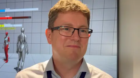 Josh Lovatt/BBC Chris Rawden is standing in front of a monitor which is displaying an animated computer-generated design. Rawden has short brown hair and is wearing black-framed glasses and a blue shirt.