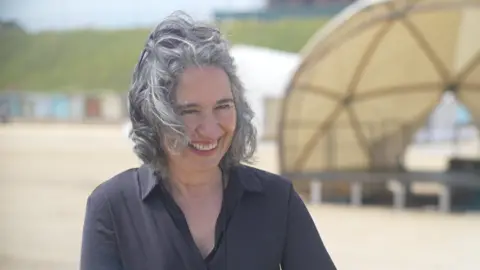 Jamie Niblock/BBC A woman with short, curly grey hair stands on a beach as a festival is beginning to be set up. She is laughing and looking away from the camera. A large tent with a stage is being erected behind her.