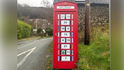 A red phone box on a grass verge next to a country road. In each window is a piece of paper with a black number on it. In place of one number, there is a piece of artwork depicting a Christmas tree