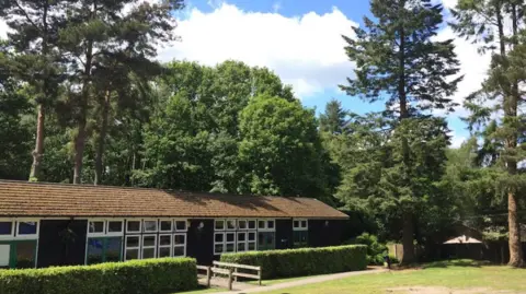 Google A single-storey building with large windows sits in a clearing surrounded by tall pine and evergreen trees. A wooden footbridge crosses a shallow drainage ditch in front of the building, with trimmed hedges running along the path. A grassed area stretches across the foreground and dense woodland rises behind the centre under a blue sky with scattered cloud.
