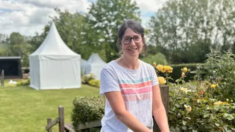 A woman with tied back brown hair and glasses is wearing a grey t-shirt with coloured stripes across the front. She is standing in front of a yellow rose bush in a garden, with a small white tent behind her. 