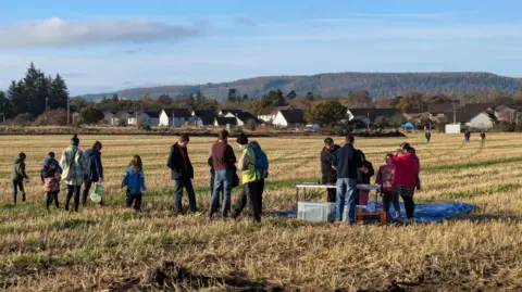 Maria de la Torre A group of people, including children, can be seen in the Smiddy Field. The grass is cut short. Houses can be seen at the edge of the field in the background.