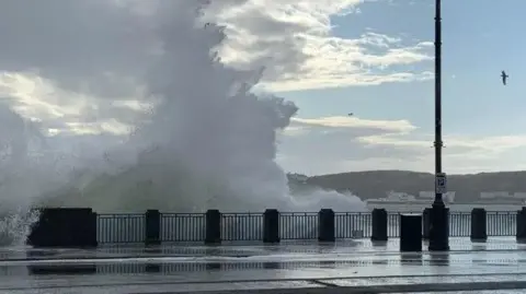 A large wave breaking over railings at the northern end of Douglas Promenade. The walkway is wet and there is a gull flying across a section of blue sky to the right.