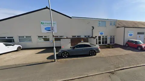 Google The former CUI International medical supplies building is a beige low rise building with a flat roof and cars parked outside.