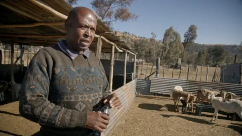 Thabo Makopo is standing by a cattle pen with some goats visible. He is wearing a patterned jumper and is holding a small water bottle.