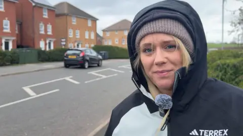 BBC A woman with blonde hair , wearing a hat with a coat up against the wind smiles at the camera. she's standing on a housing estate, with blurred out homes in the background