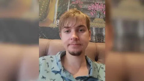 Wiltshire Council A close-up of Sean sitting on a sofa, looking straight to camera, wearing a pale blue shirt with tigers on, with a patterned wallpaper behind