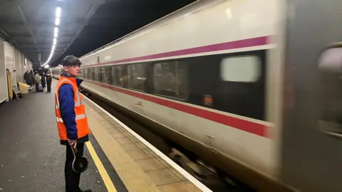 A train on a platform. An employee stands on the platform in an orange high vis vest and black trousers. There are some commuters in the background.