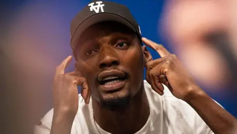 US track and field sprinter, Fred Kerley, talks with the press ahead of the Enhanced Games. He points to his face with his hands, mid-conversation.