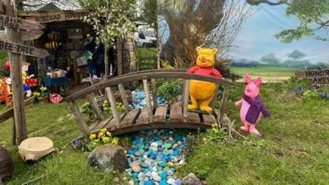 BBC/Richard Fox Winnie the Pooh and Piglet characters stand on a bridge over a stream made out of pebbles.