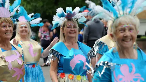 South Tyneside Council A group of women taking part in the parade. They are wearing sea-themed costumes. They are wearing blue and gold home-made tops with pictures of creatures such as crabs and dolphins on the front. They have glitter around their eyes and have blue and white feathers sticking out of their headbands.