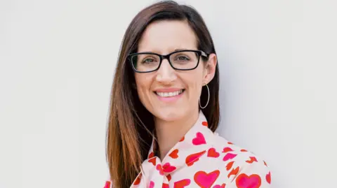 University of Aberdeen Helen Corsicadmore, who has long brown hair, glasses and a white blouse with red and pink hears, looks into the camera