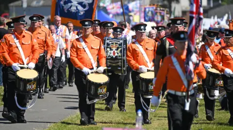 A group of people dressed in orange uniforms, wearing caps and carrying drums march in rows in Cowdenbeath in 2018.
