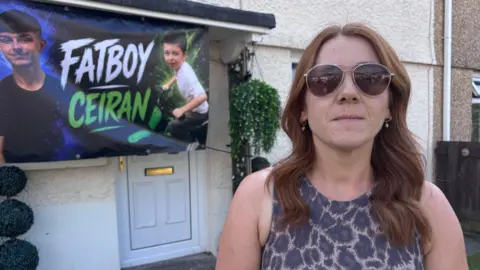 Ceiran's aunt, Becky Arnold-Jones stood outside a house There is a banner above the front door with a recent and childhood photo of Ceiran. The banner reads 'Fatboy Ceiran'.