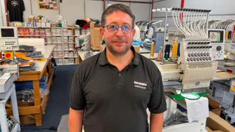 A man is standing in a black polo shirt with branding machinery behind him