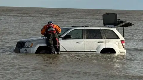A person in an orange and black jumpsuit leaning on a silver car that has water up to the top of its wheels.