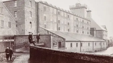 Friends of the Flaxmill Maltings A black and white photo of a large, block-shaped, brick building with two towers on its roof and a number of children in dark Victorian-style clothing standing near the front