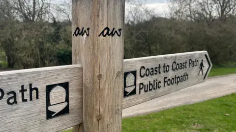 A wooden signpost stands at a junction of paths, with two horizontal arms pointing in different directions. The right‑hand arm reads “Coast to Coast Path, Public Footpath” with a walking‑figure symbol. The left‑hand arm shows part of the same acorn‑shaped National Trail marker. Trees and grassland fill the background, and a gravel path runs behind the sign.