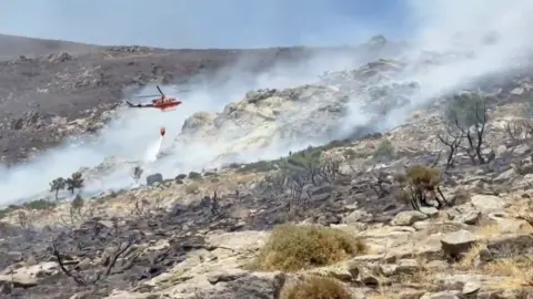 Red firefighter helicopter drops water onto a smouldering hillside.