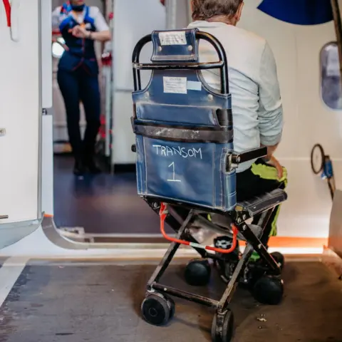 Getty Images A man sitting on a narrow chair specifically made to wheel through the aisle of an aeroplane, he is sat outside the plane waiting to board the aircraft. The chair is blue in colour and the aircrew can be seen in the background.