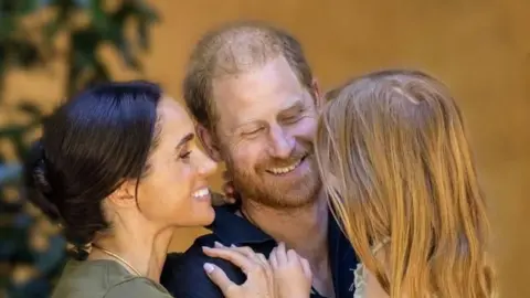 Duke and Duchess of Sussex in casual outfits embracing and holding Lilibet in a summer dress, her back is to the camera; she has long, red hair