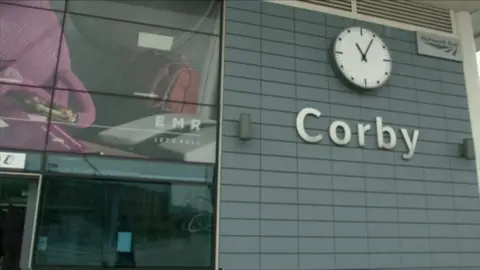 Corby Railway Station's frontage has a large sign with the town's name and a clock above that. Adverts can be seen on the left of the image