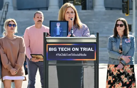 Getty Images A woman stood behind a lectern which says 'big tech on trail'. She is speaking into a small microphone and a picture of a young boy is placed on the lectern in front of her. People stand nearby with clasped hands. 