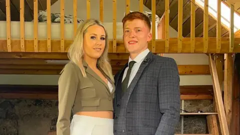 Supplied A young woman called Leighanne who has blonde hair stands facing a young man - Sam Dodd - who is wearing a suit. Behind them is a wooden balcony.
