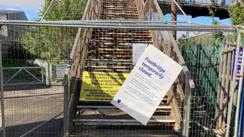 The staircase leading up to the railway footbridge is cordoned off by grey metal fencing. A sign on the fence reads: "Footbridge temporarily closed". The stair case behind it is so rusted it looks brown and orange.