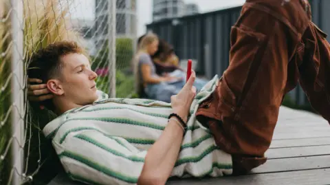 A young man lying down on the ground holding and scrolling on his smartphone.