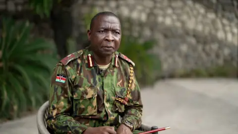 Jack Garland, BBC Godfrey Otunge, in military uniform, sits on a chair. The Kenyan is visible on the sleeve of his uniform shirt. 