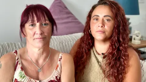 Two women with red hair sitting on a cream sofa. One has her hair tied back and is wearing a white and red vest top with a heart necklace. The other has her hair loose around her shoulders and is wearing a crew top. 