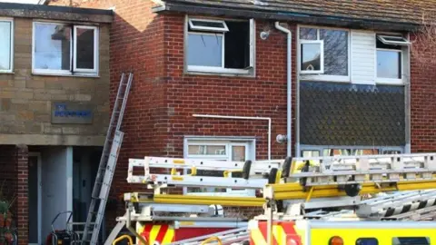 Fire engine outside two-storey flats - ladders are placed on the front of the building and windows on the second floor are open.