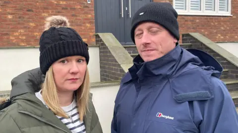 Two people wearing black woolly hats stand outside their property. Laura has a green coat and shoulder length blonde hair and is looking seriously into the camera, while her husband, wearing a blue rain jacket, has his eyes closed.