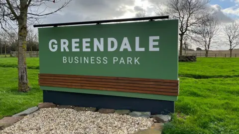 A large green and white sign for Greendale Business Park with stones at the bottom in front of the sign and grass and trees behind.