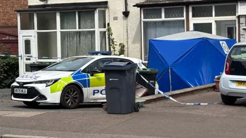 A close-up of a police car and blue West Midlands Police tent outside a semi-detached house. Police tape closes off the scene.