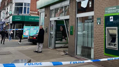 The front of a high street branch of Lloyds Bank, which has had its front entrance smashed in by a car. Blue and white police tape surrounds the front of the bank. A police officer can be seen speaking on his radio behind the cordon. Another man in a black coat is standing in front of the damaged entrance.
