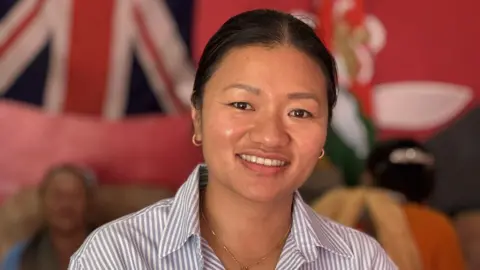 A smiling Pratikshya Gurung is standing in front of a Union Jack flag with a couple of older Gurka women sitting behind her. She's wearing an open necked blue and white striped shirt, revealing a small necklass with a cross on it.