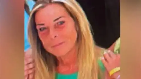 A woman with long blonde hair, aged around 60. She is wearing a green top and smiling into the camera.