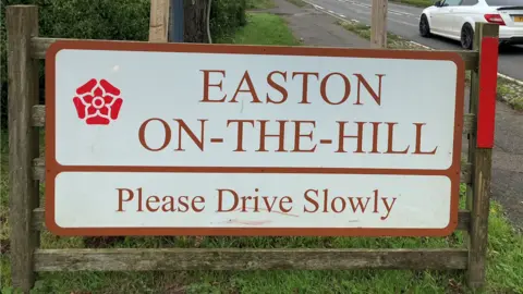 A road sign in white and red, with a rose design to the left. It is on a wooden frame, with grass around it and a white car travelling on the road to the right. 