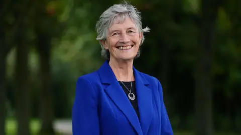 PA Media Catherine Connolly smiling. She is wearing a blue blazer, black top and a silver necklace. She has short grey hair.