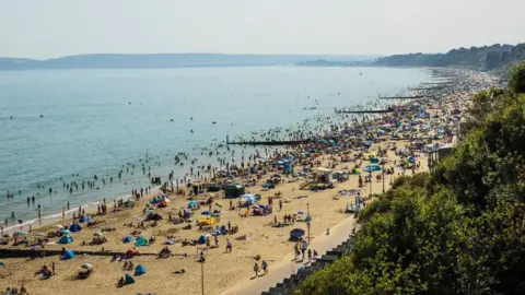 Getty Images An aerial shot of a beach on a sunny day. The beach is very busy with huge crowds of people trailing off right along the coast. Many people on the beach have tents and parasols. Hundreds can be seen paddling or swimming in the sea. 