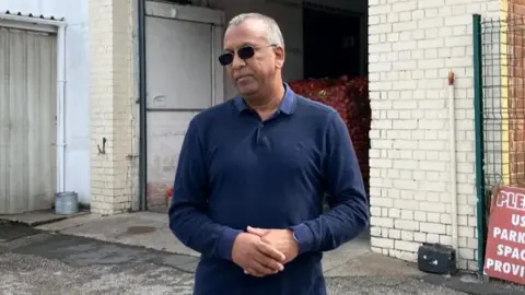 VICTORIA SCHEER/BBC Mojid Khan, owner of Sunrise Catering Supplies, stood outside his business wearing sunglasses and a blue top