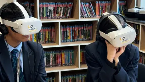 Two students, one boy and one girl sitting with virtual reality headset on. A bookshelf is behind them. They are wearing navy blazers and blue, tie with green stripes. 