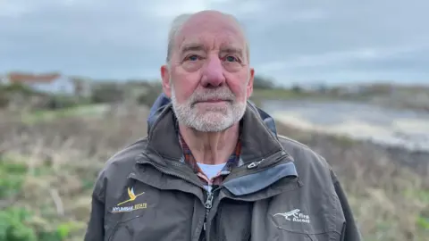 Chris has very short white hair and short white stubble. He is wearing a grey waterproof jacket.