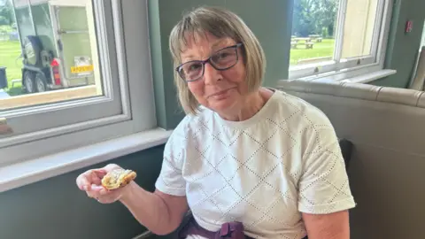 Kate Bradbrook/BBC Carol Poole is sitting in a cafe holding a piece of scone in one hand. She has a blonde bob hairdo and is wearing a white top.