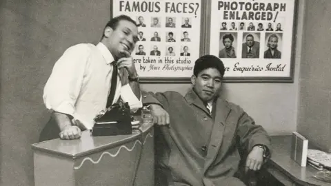 Dave Sharkey, with customer, 1962. Black‑and‑white photograph of Dave Sharkey, with customer. Dave leans against a counter with a camera placed on top, while the other sits beside the desk. Posters of sample portrait photographs hang on the wall behind them.