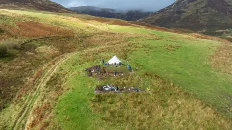 Airborne Lense/Destination Tweed An aerial image of Adie's Brae scooped settlement, showing a white teepee-style tent with two excavation pits surrounded by white tape, sitting in a hilly landscape with volunteers standing around the pits