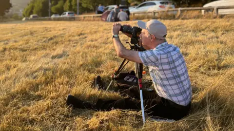 Aisha Iqbal/BBC A man in brown trousers and a checked shirt sits on dry grass holding a camera