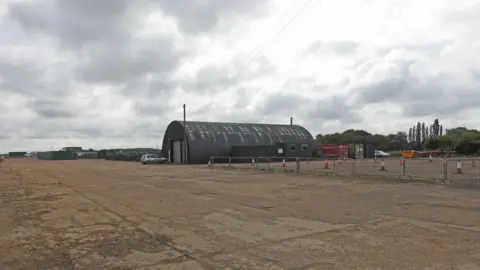 Adrian S Pye/Geograph The runway at Bourn Airfield with silver barriers and a hangar